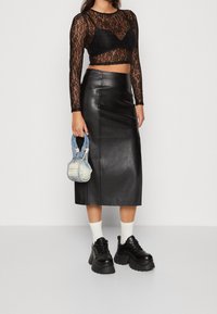 Schwarzes Spitzen-Oberteil mit langen Ärmeln, das über einem schwarzen Leder-Midirock getragen wird, kombiniert mit klobigen schwarzen Sneakers und einer kleinen Jeans-Handtasche.