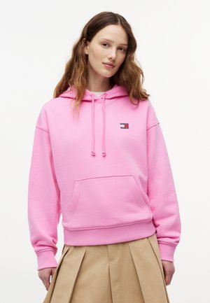 Ung kvinde med langt bølget hår iført en knaldpink hættetrøje med logo og et beige plissert skørt, stående mod en ensfarvet hvid baggrund.