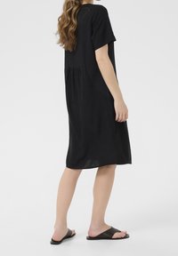 Schwarzes, kurzärmeliges Kleid mit lockerer Passform, das eine geraffte Taille, einen abgerundeten Saum und Seitentaschen aufweist. Der Stoff wirkt leicht.