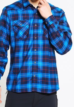 Mężczyzna w niebiesko-czarnej kraciastej koszuli na guziki z kieszeniami na piersi, poprawiający kołnierzyk prawą ręką.