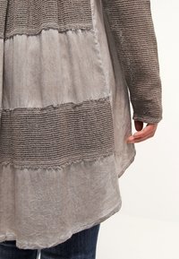 Haut beige à couches avec des détails en maille sur les manches et la taille, porté sur un jean bleu foncé, montrant une main partiellement ouverte sur le côté.