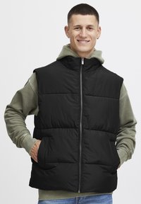 Schwarze Steppweste mit hohem Kragen, Reißverschluss vorne und zwei Fronttaschen, getragen über einem grünen Hoodie, mit glatter Textur.