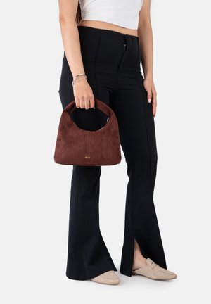 Femme portant un pantalon noir à fente sur l’ourlet et des mocassins beiges, tenant un sac à main en daim marron avec une poignée incurvée.