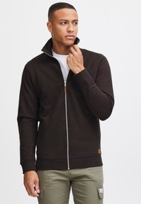 Brun zip-up-jakke med høy krage, ribbestrikkede mansjetter og hem. Mykt stoff med en glatt tekstur. Har en treknotte som detalj på glidelåsen.