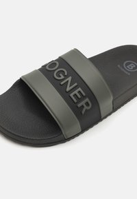 Sandal negru cu curele din cauciuc verde măsliniu. Prezintă numele mărcii embosat pe curea, un suport pentru picior texturat și un detaliu cu logo rotund.