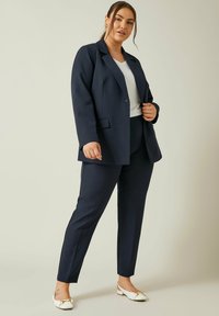 Marineblauw pak met een op maat gemaakte blazer en slim-fit broek; gecombineerd met een witte top en witte schoenen met een gouden accent.