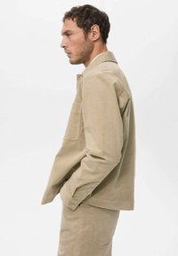 Beige corduroy jacket with a collared neckline, single chest pocket, and long sleeves. Model wears matching beige pants.