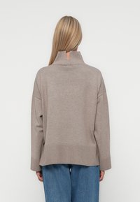 Light brown, ribbed sweater with a high collar and drop shoulders, featuring a relaxed fit and split hem, worn over blue wide-leg jeans.