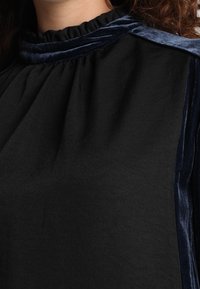 Nahaufnahme einer Person, die ein schwarzes Oberteil mit dunkelblauem Samtbesatz am Ausschnitt und den Schultern trägt.