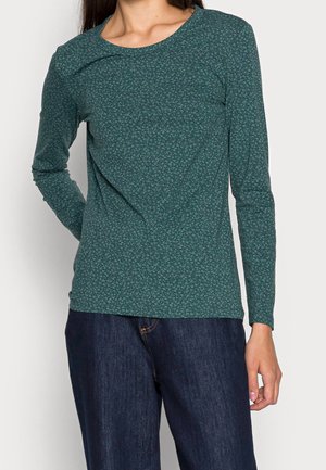 Femme portant une chemise à manches longues bleu sarcelle avec un petit motif floral et un jean bleu foncé, debout avec une main dans le dos.