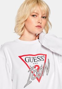 Sweatshirt blanc arborant un grand logo triangle rouge avec "GUESS" et "ICON" en paillettes argentées, sur un fond uni.