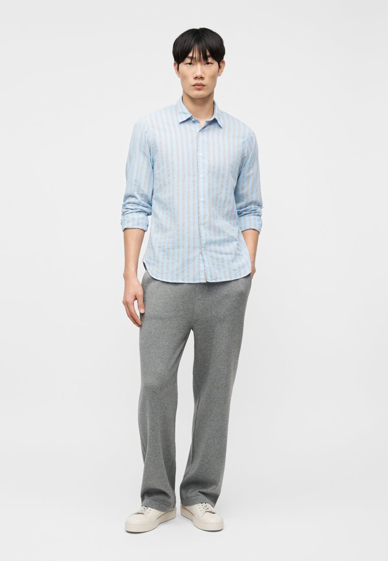 Young man standing with hands in pockets, wearing a blue striped button-up shirt, gray pants, and white sneakers on a white background.