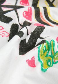 Tissu blanc avec des imprimés colorés et texturés incluant des cœurs rouges, des formes noires et des motifs abstraits vert-jaune.