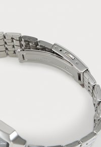 Montre en métal argenté avec un bracelet à maillons. Dispose d'une fermeture à clip et d'une texture lisse et polie. Style minimaliste sans détails visibles.