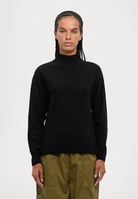 Pullover nero a maglia con colletto alto e maniche lunghe, caratterizzato da un orlo a coste. Indossato con pantaloni cargo verde oliva.