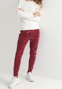 Femme croisant les bras, vêtue d'un pull en maille blanche, d'un pantalon en velours côtelé bordeaux et de baskets blanches, debout devant un fond clair uni.