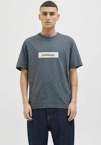 Grå bomulds-T-shirt med et rektangulært hvidt mærke med "Jack & Jones" i sort tekst, parret med løse mørke jeans.