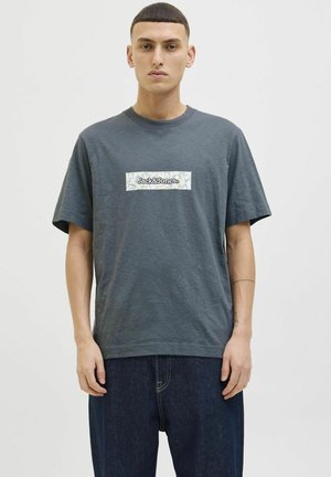 Grå bomulds-T-shirt med et rektangulært hvidt mærke med "Jack & Jones" i sort tekst, parret med løse mørke jeans.