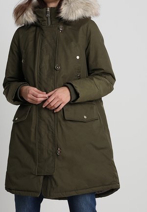 Femme portant un parka d'hiver vert olive avec capuche bordée de fourrure, fermeture à boutons et à glissière, poches à rabat, et un jean bleu.