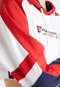 Felpa con maniche a blocchi di colore rosso, bianco e blu navy, con dettagli in rete e parte del logo Tommy Sport sul petto, indossata da una persona.