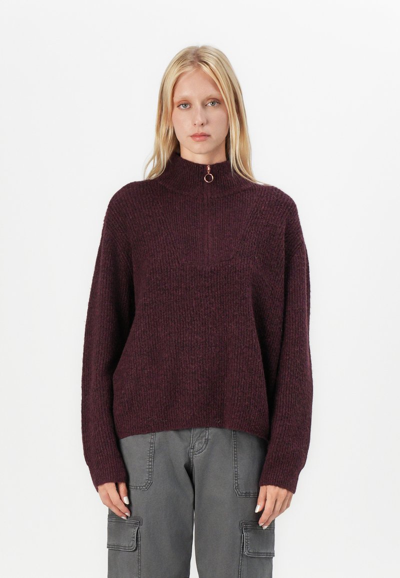 Pull en tricot bordeaux avec un col haut et une fermeture éclair quart. Présente un tissu texturé et des épaules tombantes pour un ajustement décontracté.