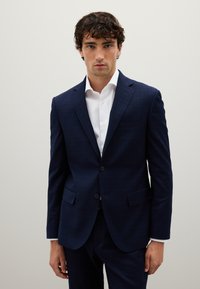 Blazer a quadri blu navy realizzato in un tessuto con una texture liscia. Caratterizzato da due bottoni, rever a punta e una tasca sul petto. Indossato sopra una camicia bianca.