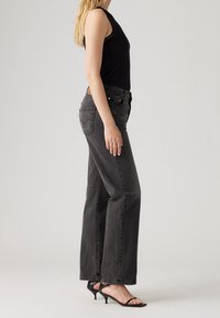 Mujer vestida con una blusa sin mangas negra, jeans de pierna recta de talle alto en gris oscuro y sandalias de tacón de aguja negras con tiras, vista lateral.