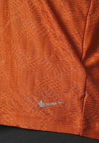 Haut en tissu texturé orange avec un logo Nike en argent scintillant, mettant en avant des motifs complexes et une finition lisse.