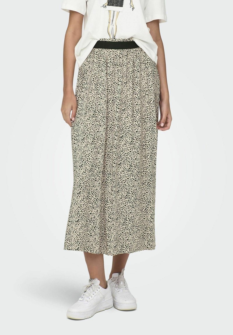 Jupe maxi de couleur beige clair avec un motif tacheté noir. Taille élastique, tissu plissé, portée avec des baskets blanches.
