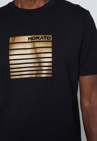 Black t-shirt featuring gold horizontal stripes and the word "MORATO" above the design. Made of smooth fabric with a crew neckline.