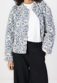 Veste matelassée avec un motif paisley bleu et blanc, dotée d'un col rond, d'une ouverture sur le devant et de deux poches avant. Associée à un haut blanc et un pantalon noir.