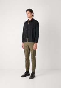 Camisa negra de botón con dos bolsillos en el pecho, combinada con pantalones cargo verde oliva que tienen bolsillos laterales, y zapatos negros.