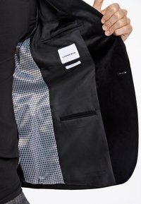 Zwarte jasvoering met een gladde stof, voorzien van een grijs patroon aan de binnenkant, een binnenzak en een merklabel. Accenten zijn onder andere glanzende hardware.