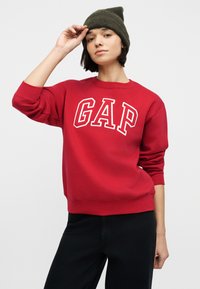 Νεαρή γυναίκα που φοράει κόκκινο φούτερ GAP και πράσινο ελιάς σκούφο, σηκώνει ελαφρώς τον σκούφο με το δεξί της χέρι σε απλό φόντο.