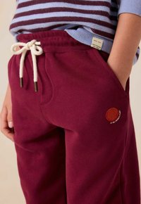 Burgunderrote Jogginghose mit einem Kordelzugbund, ausgestattet mit einem kleinen, gestickten orangenen Kreis mit Text. Hergestellt aus weichem Stoff.