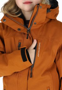 Orange wasserdichte Jacke mit hohem Kragen, Reißverschluss und Druckknopf-Verschlüssen, elastischen Bündchen und Logodetail auf der Vorderseite. Weicher Stofftextur.