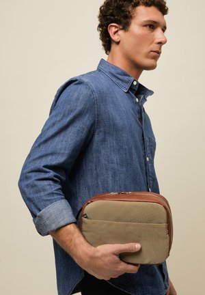 Muškarac u plavoj teksas košulji drži smeđu i bež torbicu s patentnim zatvaračem na neutralnoj pozadini, okrenut udesno.