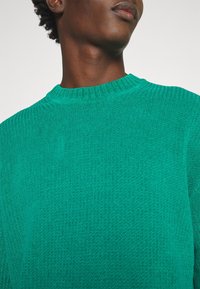 Klargrön stickad tröja med ribbad textur och rundad halsringning, som framhäver mjukt tyg och en figurnära silhuett.