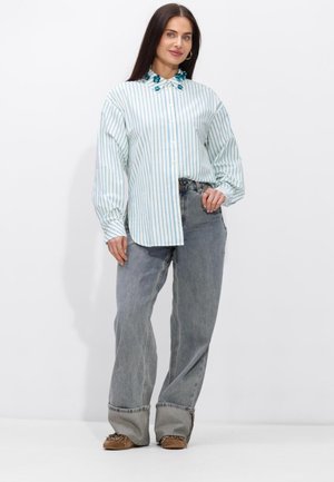 Vrouw met lang donker haar, draagt een losse blauw gestreepte blouse met knopen en een bloemenkraagdetail, wijde grijze jeans en bruine loafers.