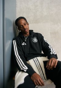 Czarna bluza z kapturem z białymi paskami Adidas, z logo i trzema gwiazdkami. Model siedzi na teksturowanym białym i czarnym kocu przypominającym futro.