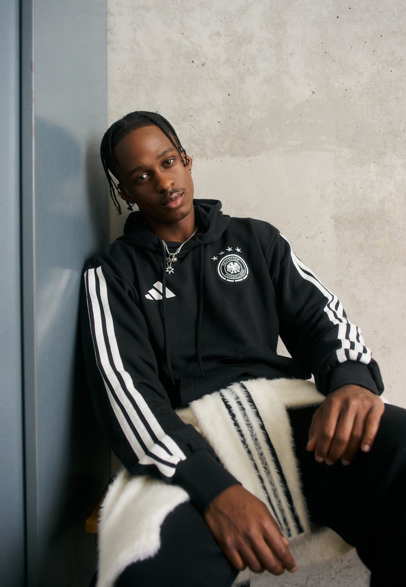 Czarna bluza z kapturem z białymi paskami Adidas, z logo i trzema gwiazdkami. Model siedzi na teksturowanym białym i czarnym kocu przypominającym futro.