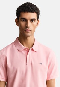 Muž s krátkymi kučeravými vlasmi, ktorý nosí svetloružové polo tričko s golierom a dvoma gombíkmi, čelí dopredu na jednofarebnom svetlom pozadí.