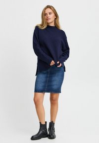 Marineblauer Strickpullover mit hohem Kragen, kombiniert mit einem Jeans-Bleistiftrock und schwarzen Knöchelbooties mit Schnallenakzenten.