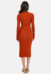 Geripptes, langärmeliges Kleid in tiefem Orange, figurbetontes Design mit Taillengürtel, reicht bis zur Mitte der Wade, strukturierter Stoff, kombiniert mit hellbraunen Absätzen.