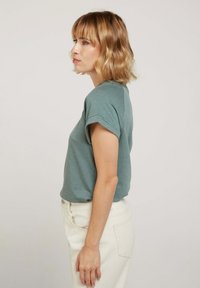 T-shirt turquoise à manches courtes avec des poignets retroussés, fabriqué en tissu doux. Porté avec un jean blanc taille haute, vue de profil.