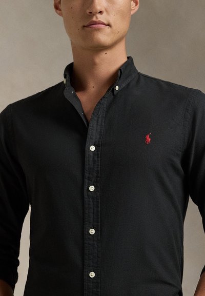 Moški, oblečen v črno srajco z gumbi in rdečim izvezenim logotipom polo igralca na levi strani prsnega koša, na nevtralnem ozadju.