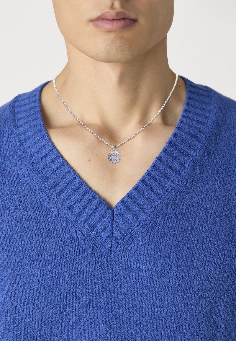 Pull en V bleu texturé avec un col côtelé, associé à un collier en chaîne argenté avec un pendentif en forme de coquillage.