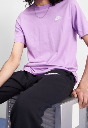 Persoon draagt een paarse Nike t-shirt en zwarte Nike trainingsbroek, zit op een metalen kist, hand rust op de rand.