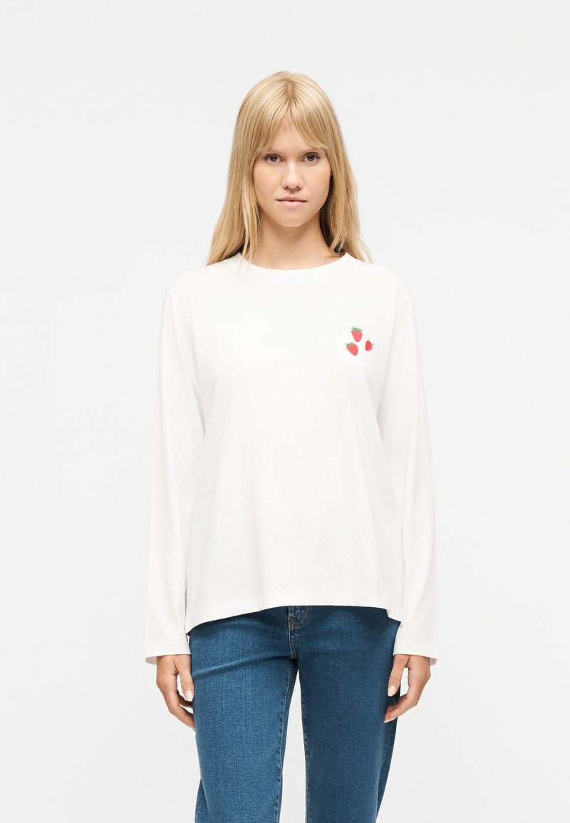 Γυναίκα με μακριά ξανθά μαλλιά, ντυμένη με άσπρο πουκάμισο μακρυμάνικο με τρεις μικρές φράουλες στο στήθος και μπλε τζιν, στέκεται μπροστά σε λευκό φόντο.