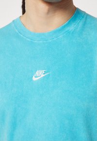 T-shirt Nike turquoise avec col rond, fabriqué en tissu doux. Présence d'un logo brodé blanc sur la poitrine et de détails de couture visibles.